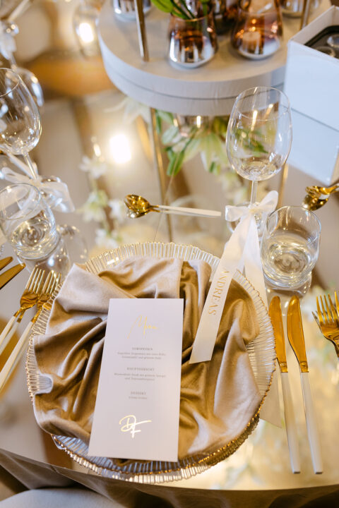 Biała karta menu na złotej serwetce, złote sztućce i kieliszek z personalizowaną wstążką na weselu Dani i Franka